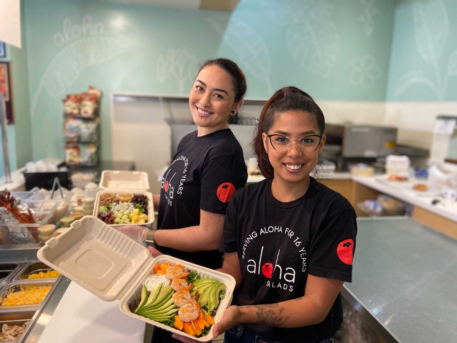 Two women at a salad bar, holding open containers of fresh salads, smiling at the camera.